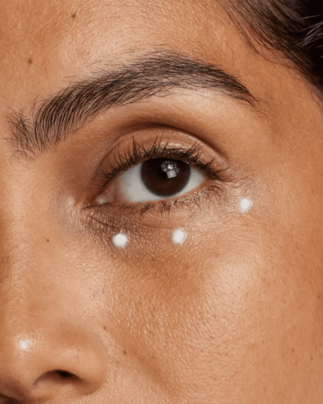 Close-up of an Indian woman with three dots of Agency Skincare Eye Formula moisturizer under her eye, demonstrating targeted application for the fine lines, puffiness, and dark circles.