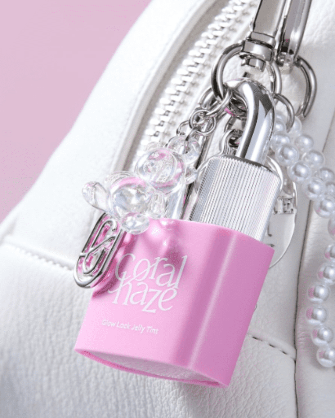 White purse close-up clipped with Coral haze Glow Lock Jelly Tint as a charm, surrounded by a chain of pearls and a clear bear accessory on a faded pink-purple background, illustrating a stylish, on-the-go self-love practice.