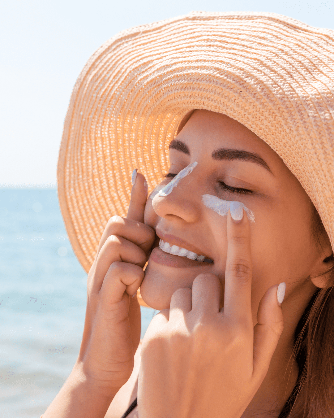 Woman in sun hat applying acne safe sunscreen under her eyes while smiling at the beach