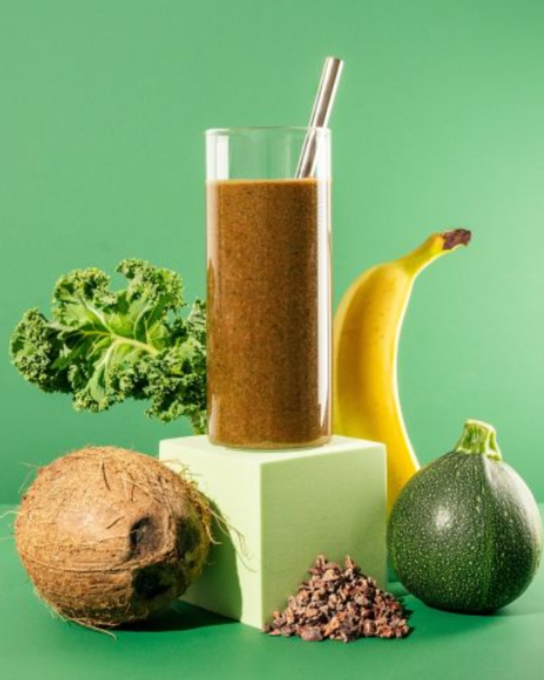 Glass of SmoothieBox Morning Cocoa smoothie with a straw, styled on a green background with kale, banana, coconut, cocoa pieces, and zucchini. The setup highlights fresh, nutrient-rich ingredients of the blend. Drink this as a self-love practice.
