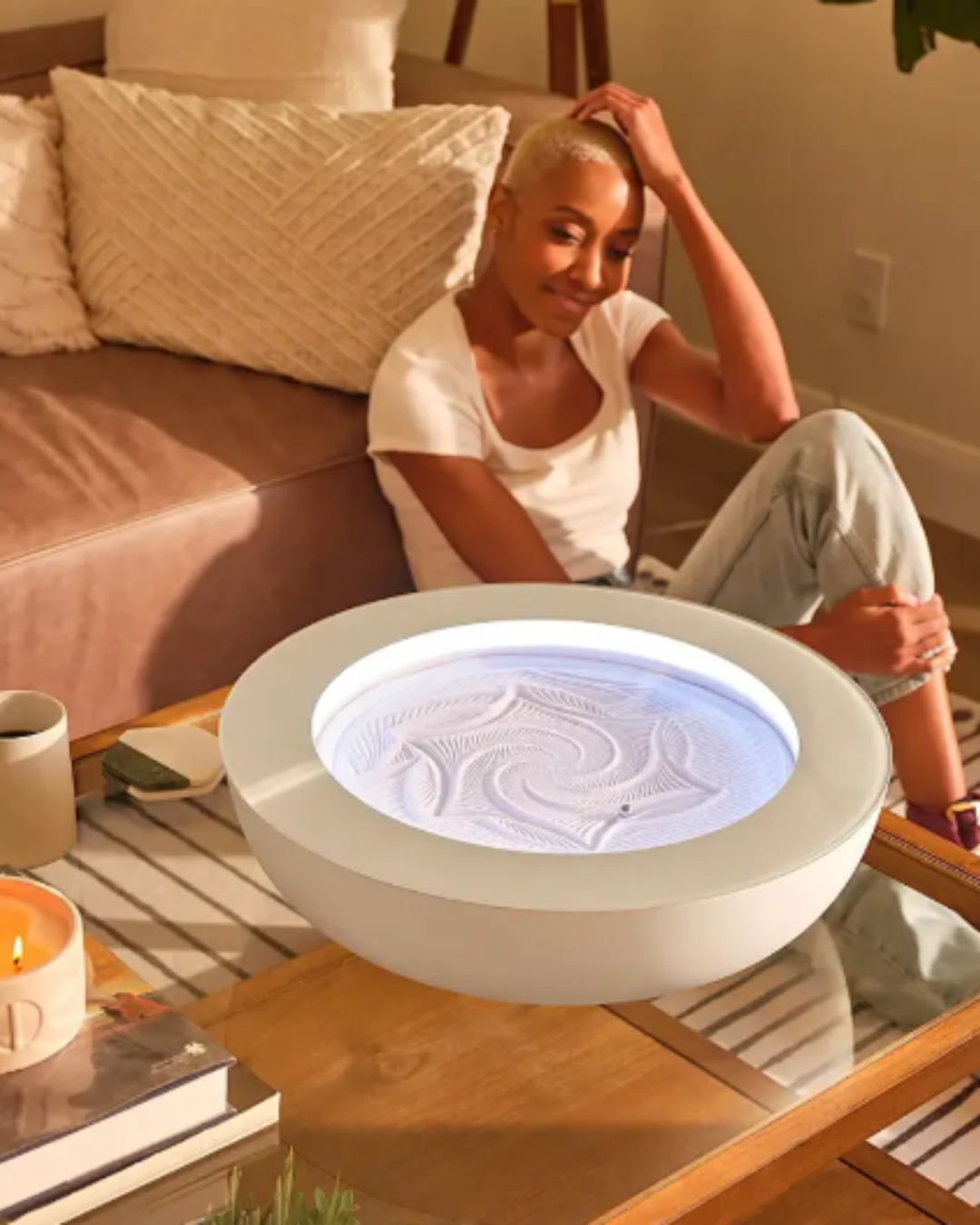 Black woman in white T-shirt and sweatpants admiring HOMEDICS Drift Meditation Sand Table on a living room table in front of a white coach, enhancing her home self-love practice.