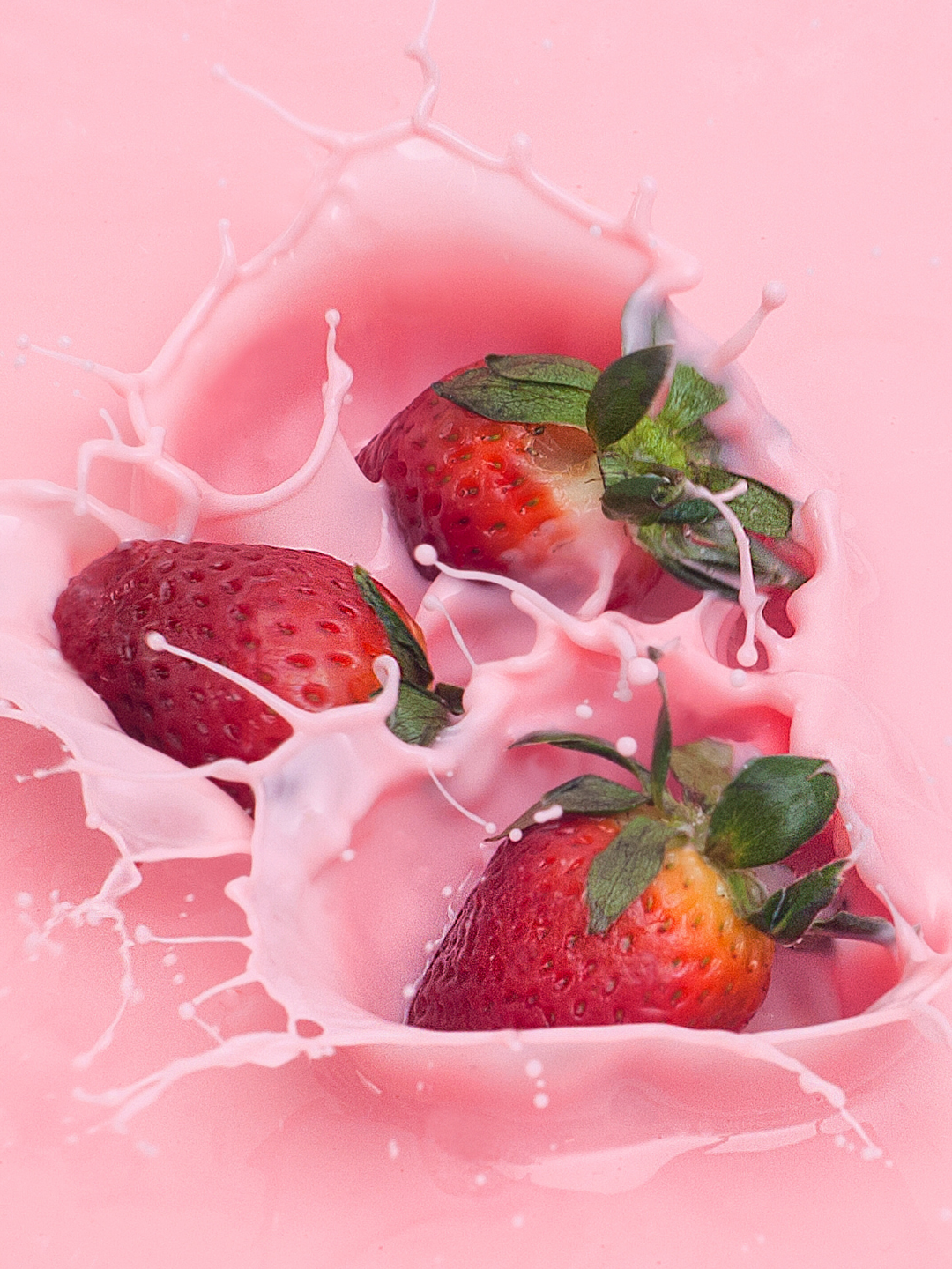 Three red strawberries with green stems falling into bright pink liquid, creating a dramatic splash, representing fruity notes in the candle.