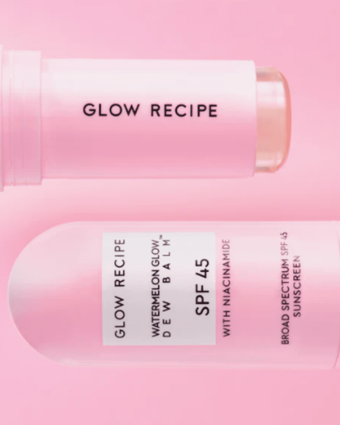 Two Glow Recipe Watermelon Glow Niacinamide Dew Balm SPF 45 products presented side by side and sideways on a pink background with white bursts of light behind the products, one with the cap off showing the clear stick, emphasizing acne friendly sun care.