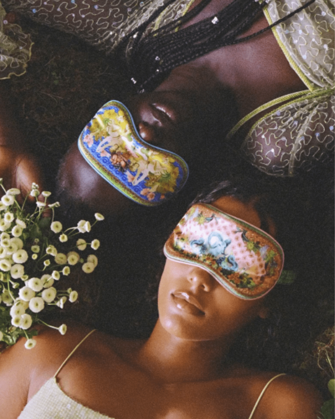 Two women lying on a bed of moss with white flowers nearby; one is upside down and the other is lying normally, both wearing the Slip Pure Silk Zodiac Sleep Mask, one in a light tank top, highlighting comfort and restful sleep.