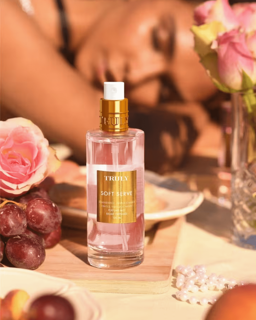 Truly Soft Serve Perfume Mist displayed on a table with grapes, flowers, and pearls, with a relaxed, brown-skinned woman in the background, highlighting this playful ice cream perfume.