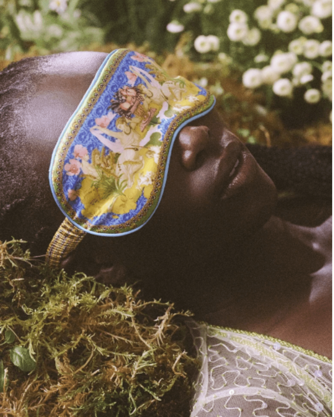 Black woman sleeping on a bed of moss with white flowers in the background, wearing a white lace top and a Slip Pure Silk Zodiac Sleep Mask showcasing luxurious nighttime relaxation.