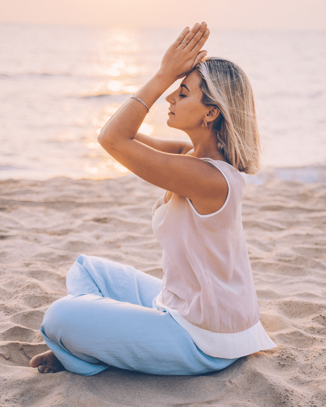 young blonde woman in jeans sitting on the beach meditating representing people wearing acne safe sunscreen.