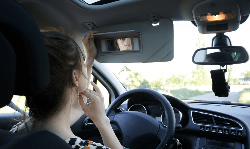 Example of using To Go Spa products in your car; white woman with brown hair looking at herself in a visor mirror inside her care, outdoors.