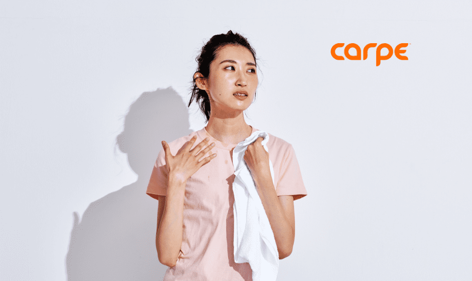 Asian woman holding a white towel in her right hand, left hand on her shoulder, looking sweaty with hair in a ponytail, wearing a pink shirt, white background and shadow behind her, Carpe logo visible.
