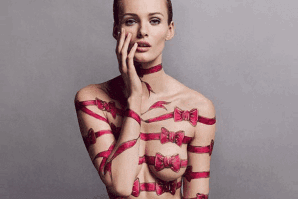 Woman with hand-painted pink bows on her naked body, sitting cross-legged with BONBON Eau de Parfum bottle in her lap, highlighting the bold candy perfume.