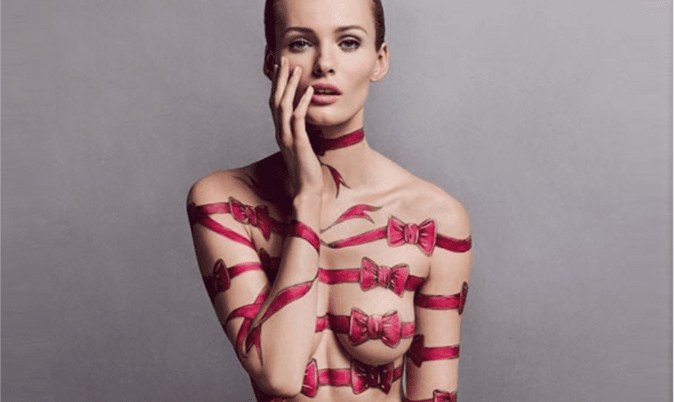 Woman with hand-painted pink bows on her naked body, sitting cross-legged with BONBON Eau de Parfum bottle in her lap, highlighting the bold candy perfume.