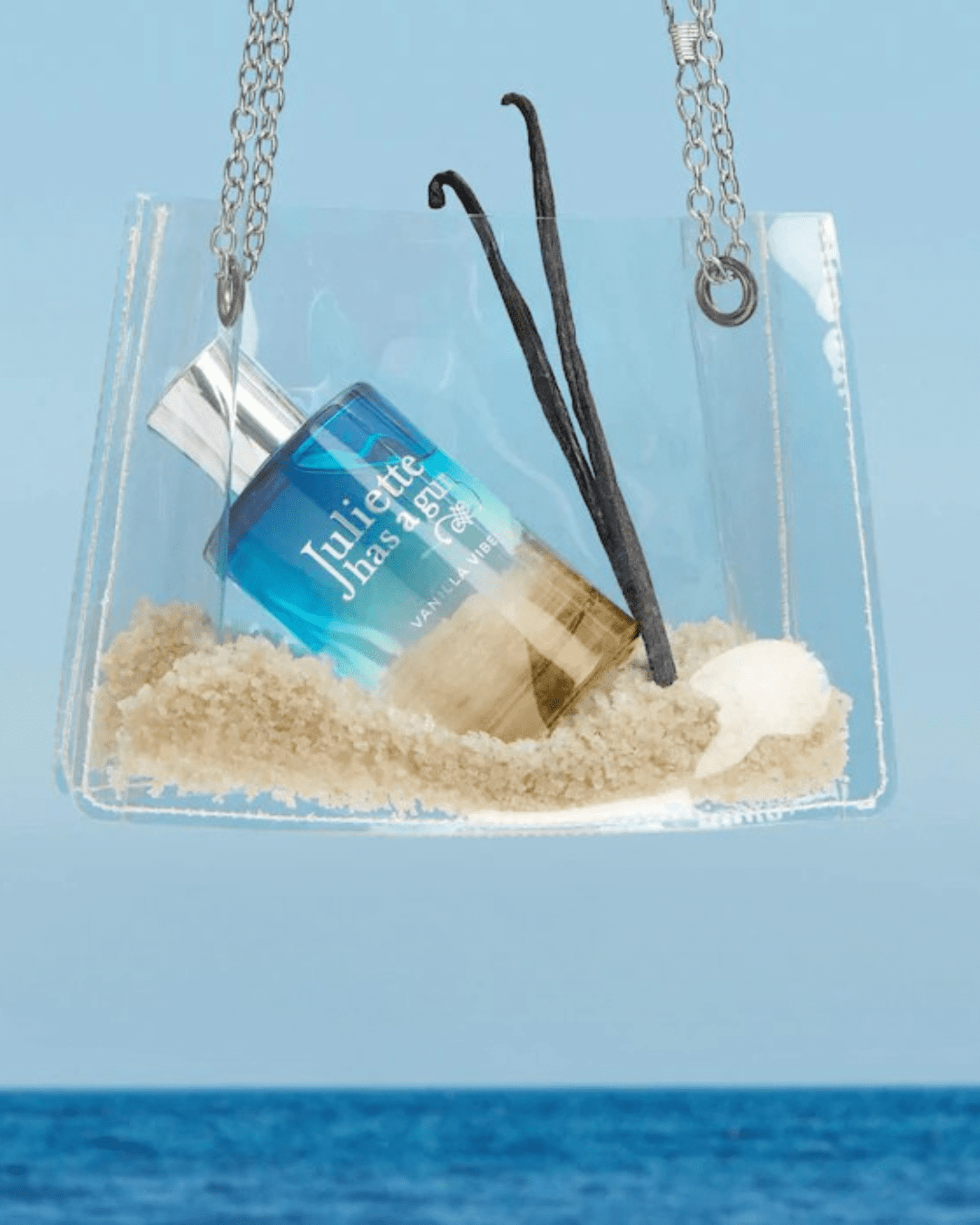 Juliette Has a Gun Vanilla Vibes nestled in a clear bag with a silver chain against a calm ocean horizon, surrounded by sand and vanilla beans, ideal for layering with ice cream perfumes.