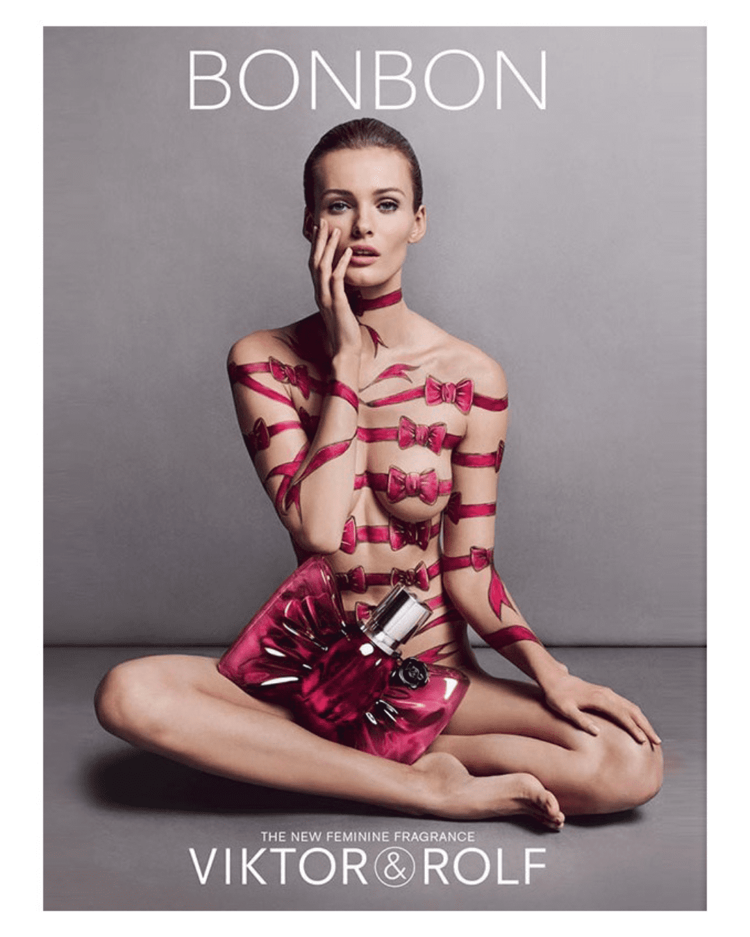 Woman with hand-painted pink bows on her naked body, sitting cross-legged with BONBON Eau de Parfum bottle in her lap, highlighting the bold candy perfume.