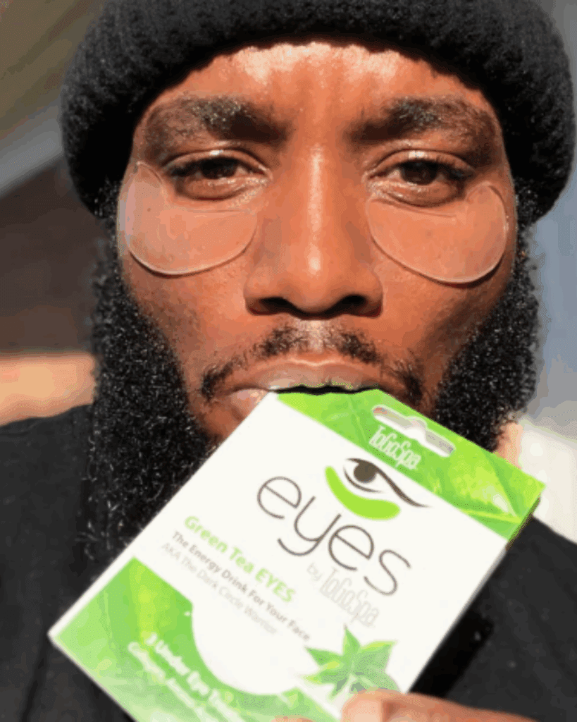 Black man wearing a black beanie and has a beard, wearing To Go Spa Green Tea Under Eye Masks with the product package held in his mouth.