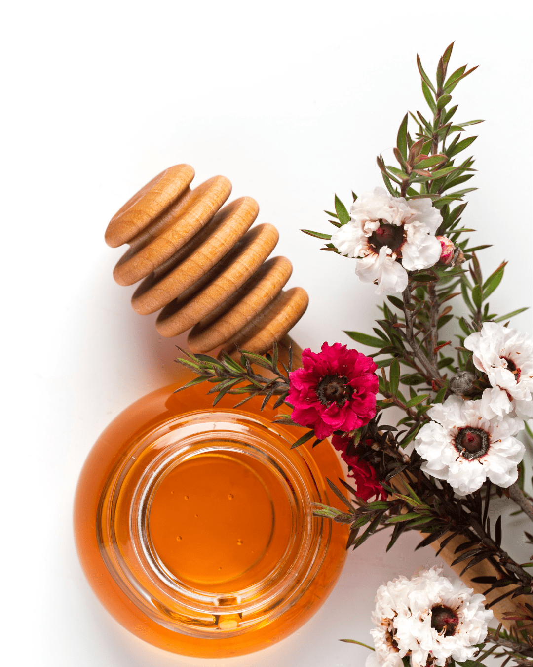 Small jar of honey with honey dipper, greenery, pink flower, and white blossoms on a white flat surface.