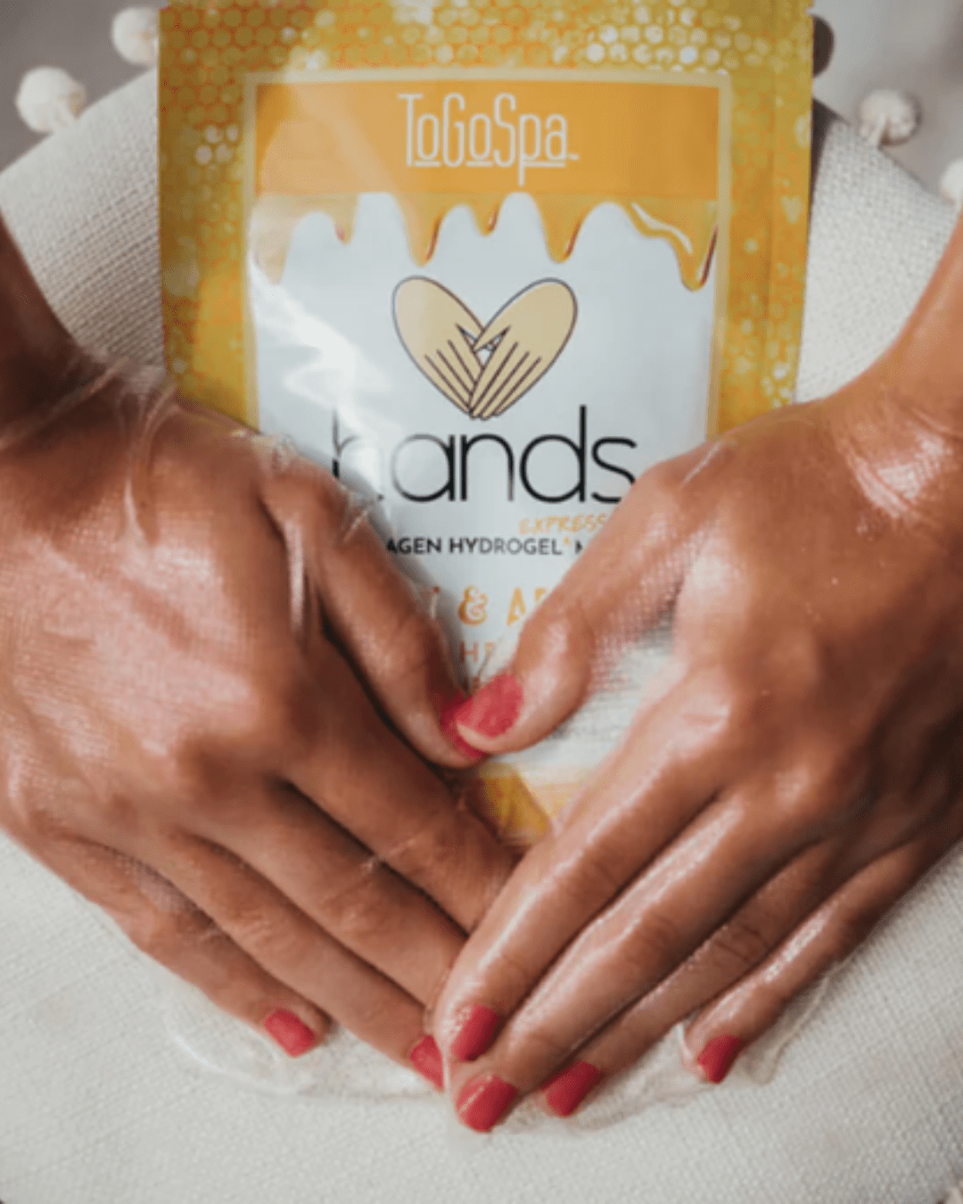 Woman with red nail polish wearing To Go Spa Honey + Arnica HAND Mask, hands resting on white pillow with decorative tassels.