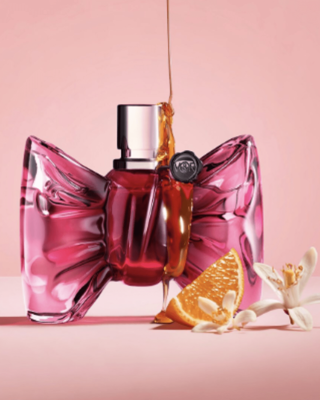 BONBON Eau de Parfum bottle on a pink surface with orange slice, jasmine, and honey dripping from above onto the perfume bottle, emphasizing the sweet candy perfume essence.