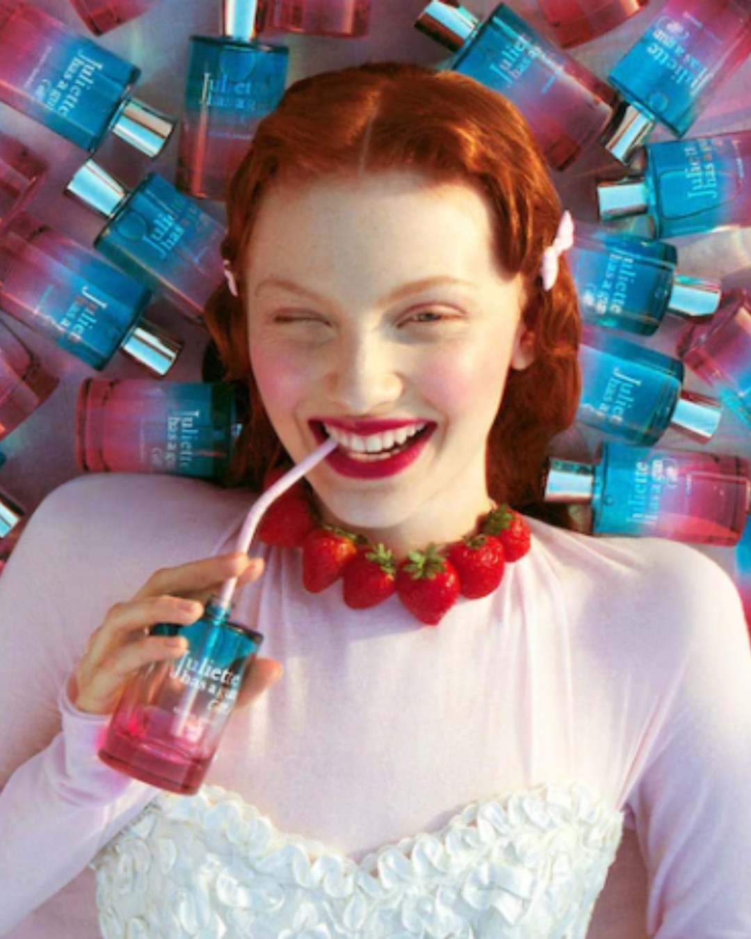 Redheaded woman with bright pink lipstick, a strawberry necklace, and a pink long sleeved shirt and white floral corset lying on multiple Juliet Has a Gun Miami Shake bottles, emphasizing the playful and fruity ice cream perfume.