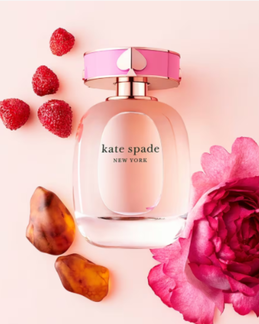 Kate Spade New York Eau de Parfum bottle lying flat on a soft pink background with a pink rose, fresh raspberries, and warm woody notes, showcasing strawberry perfumes.