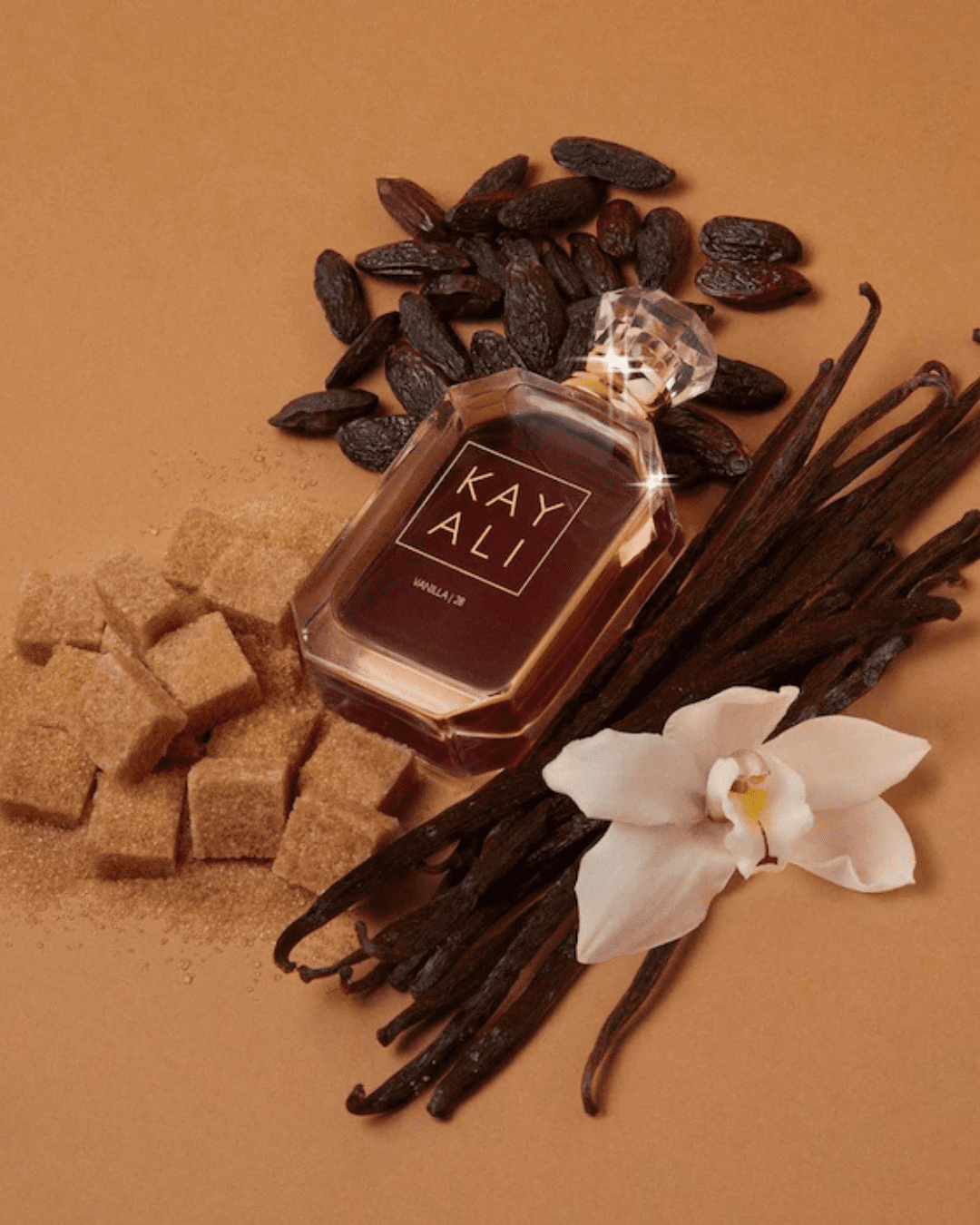 Kayali Vanilla 28 perfume bottle resting on a tan surface, surrounded by salted caramel, almonds, butter tonka accords, and a white flower, perfect for layering with ice cream perfumes.
