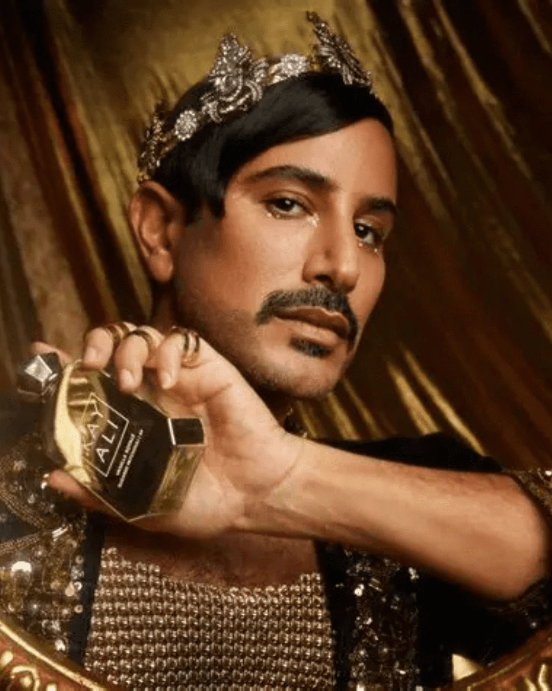Man wearing a silver crown, black jacket with gold sequins, and subtle facial hair, holds Kayali Vanilla Royale Sugared Patchouli | 64, a Crème Brûlée inspired perfume in front of a gold curtain.
