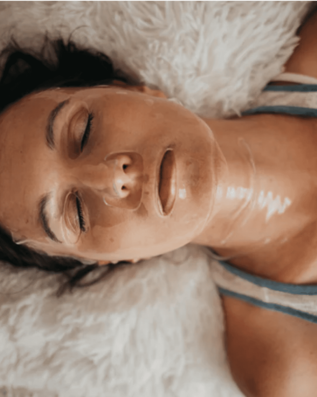 White woman with brown hair lying on a fuzzy white pillow, wearing To Go Spa Face & Neck Ice Water Mask, wearing a tank top and relaxing with eyes closed.