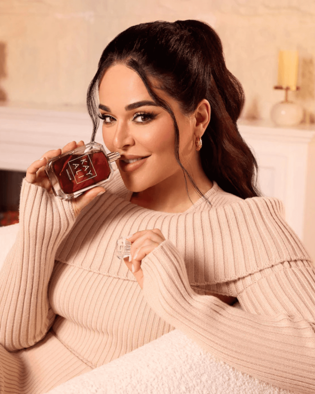 Mona Kattan, founder of Kayali, smelling Kayali Vanilla 28 perfume while wearing a cream sweater on a white couch with a fireplace behind her, perfect for layering with ice cream perfumes.