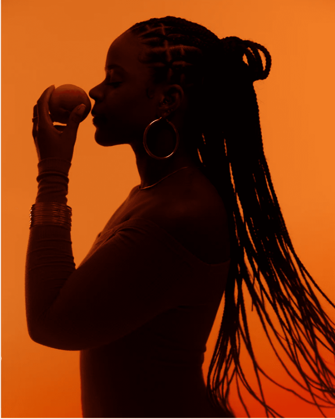 Fragrance influencer Jade Bodley posing in a shadowed setting with an illuminated orange backdrop, promoting her collaboration on the Le Monde Gourmand Pêche Délicieuse Eau de Parfum.