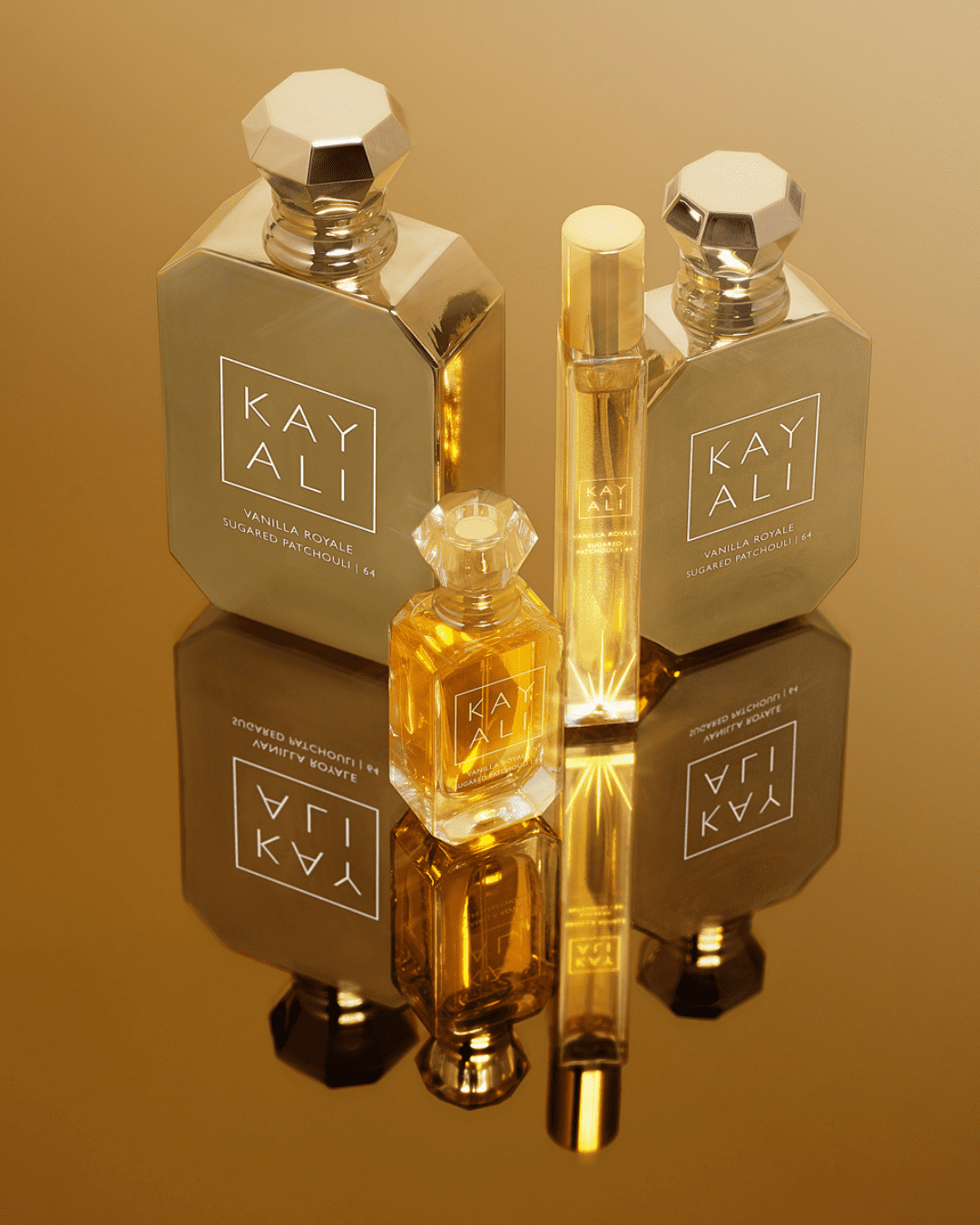 Kayali Vanilla Royale Sugared Patchouli | 64 perfume bottle in multiple sizes on a mirrored surface against a caramel-brown background, showcasing the Crème Brûlée inspired scent.