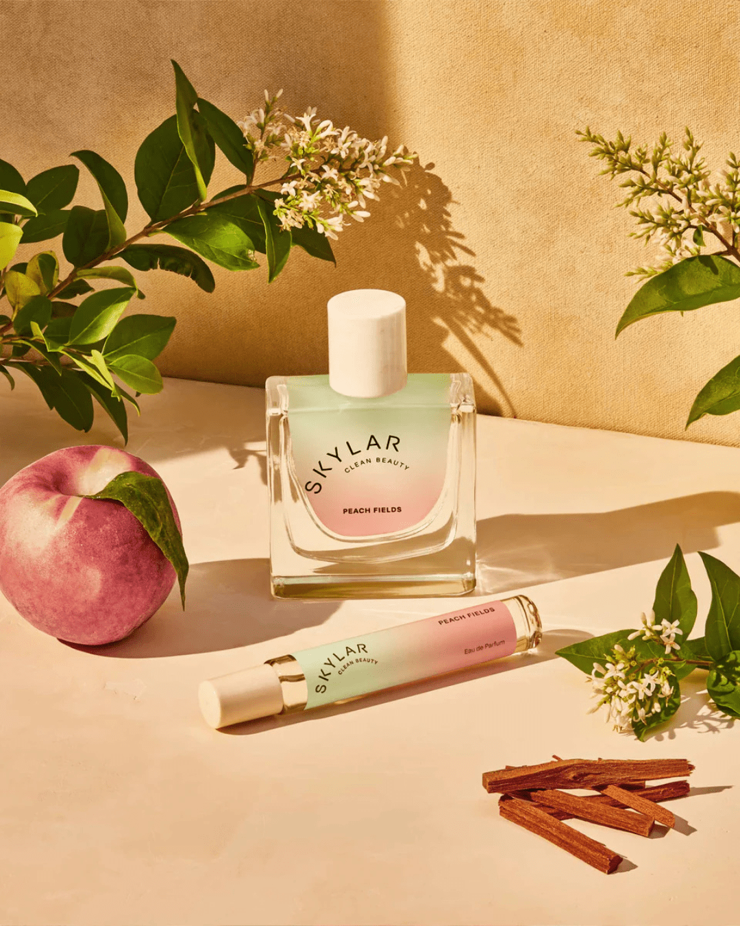 Skylar Peach Fields Perfume displayed with travel-sized bottle, jasmine, and a peach on a sandy cream surface, perfect for layering with creamy ice cream perfumes.
