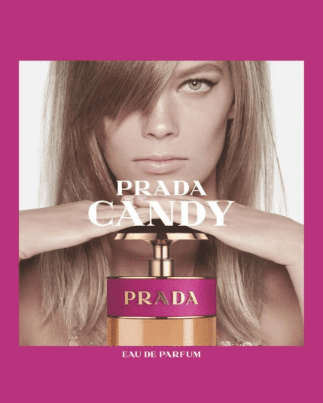 Blonde woman resting chin on hands atop a large Candy Eau de Parfum bottle, looking at the camera with a dark pink background.