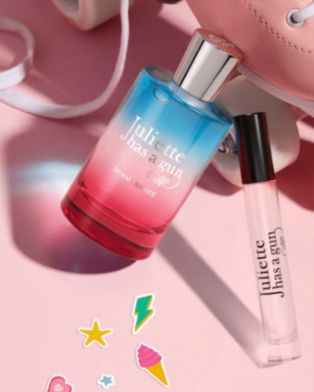 Miami Shake by Juliette Has a Gun leaning against a pink roller skate with fun stickers, next to a travel-size bottle, showcasing this playful ice cream perfume.