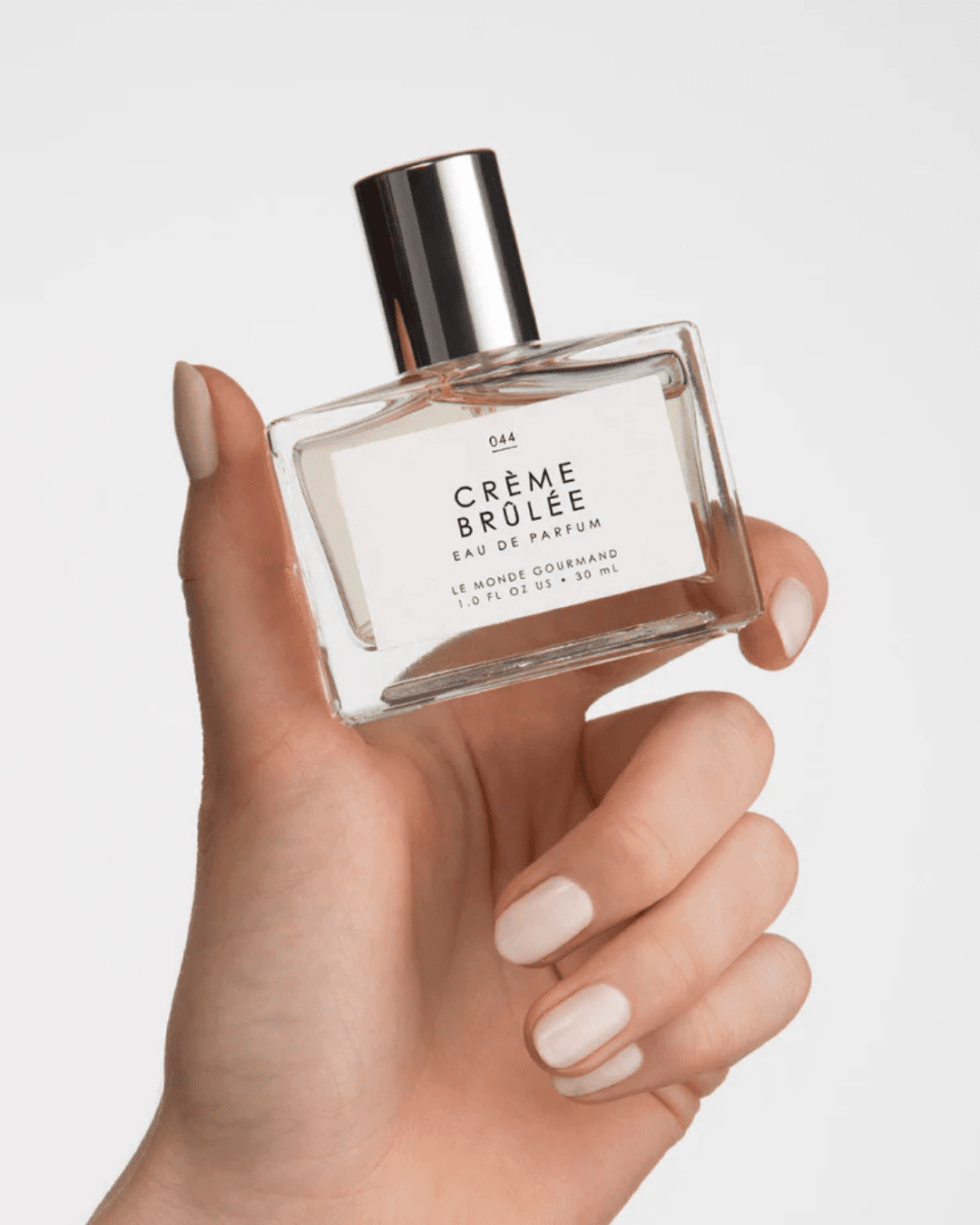 Hand holding Le Monde Gourmand Crème Brûlée Eau de Parfum bottle against a white background, highlighting the luxurious dessert-inspired fragrance.