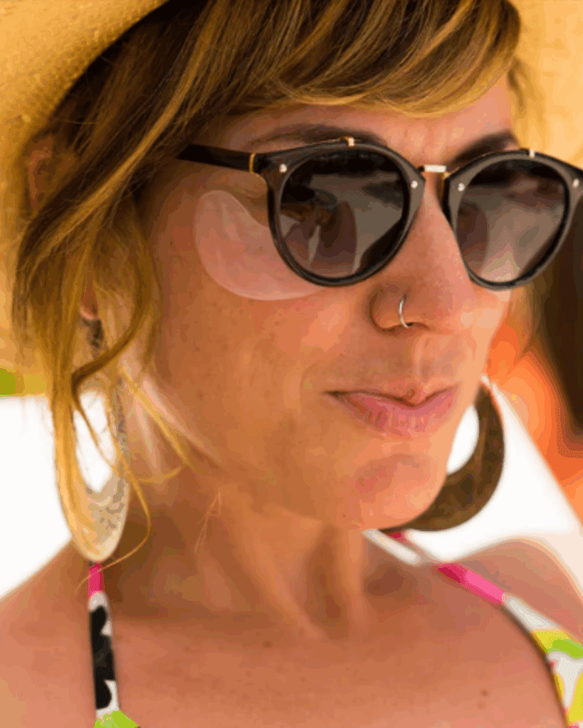 Woman in a sun hat, sunglasses, bikini, and gold earrings, wearing To Go Spa Hangover Ice Water Under Eye Patches outdoors.