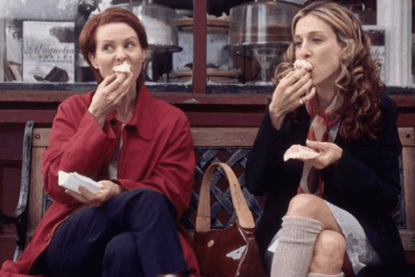 Sex and the City characters Cynthia and Carrie Bradshaw enjoying cupcakes in front of a cupcake shop, evoking cupcake perfume vibes.