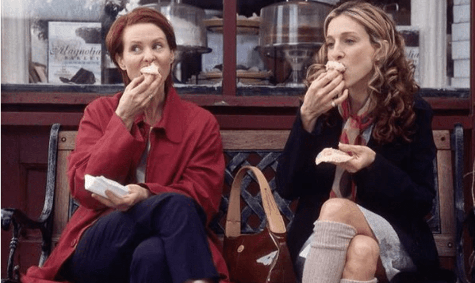 Sex and the City characters Cynthia and Carrie Bradshaw enjoying cupcakes in front of a cupcake shop, evoking cupcake perfume vibes.