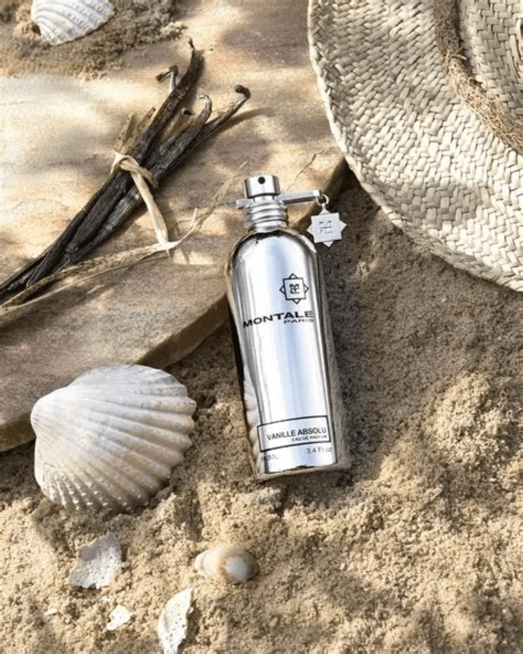 Vanille Absolu by Montale Paris perfume bottle leaning against a stone circle with vanilla beans on top, seashells, a large shell, and a straw beach hat on sand, styled to evoke the sweetness of cupcake-inspired perfumes.