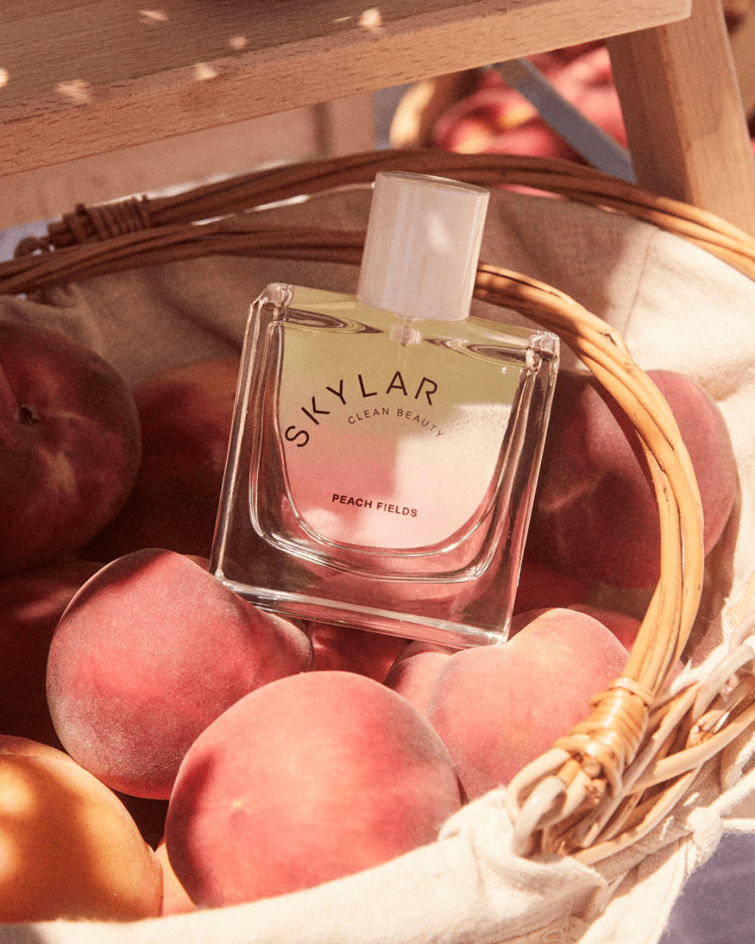 Skylar Peach Fields Perfume bottle nestled in a cotton fabric-lined basket of peaches, a sweet and fruity fragrance ideal for layering with ice cream perfumes.