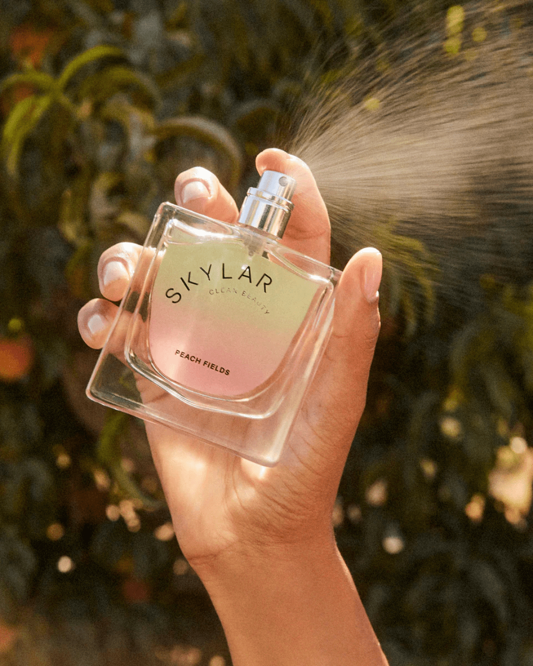 Hand spraying Skylar Peach Fields Perfume with leaf botanical background, a fresh fruity scent perfect for layering with ice cream perfumes.