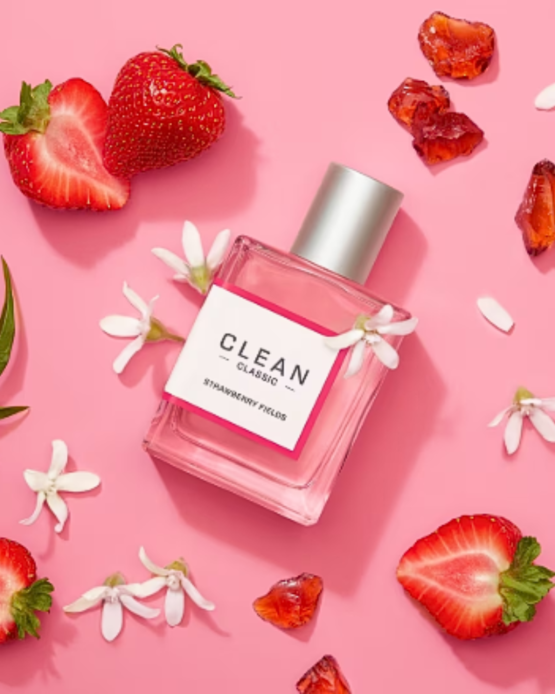 Clean Classic Strawberry Fields Eau de Parfum bottle laying at an angle on a pink background, surrounded by jasmine flowers, showcasing the beauty of strawberry perfumes.