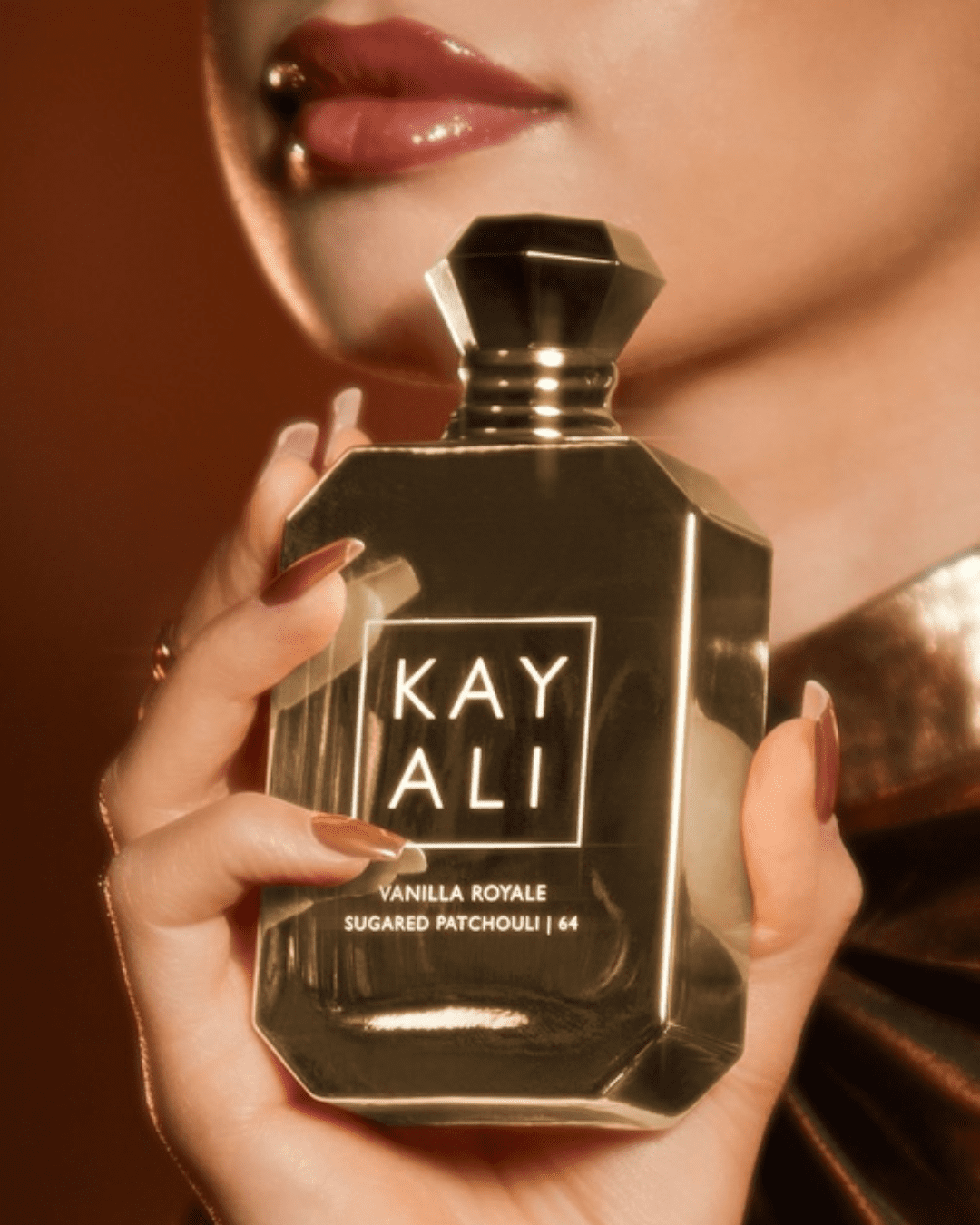 Woman wearing a gold shirt and muted pink lipstick holds Kayali Vanilla Royale Sugared Patchouli | 64, a Crème Brûlée inspired perfume.