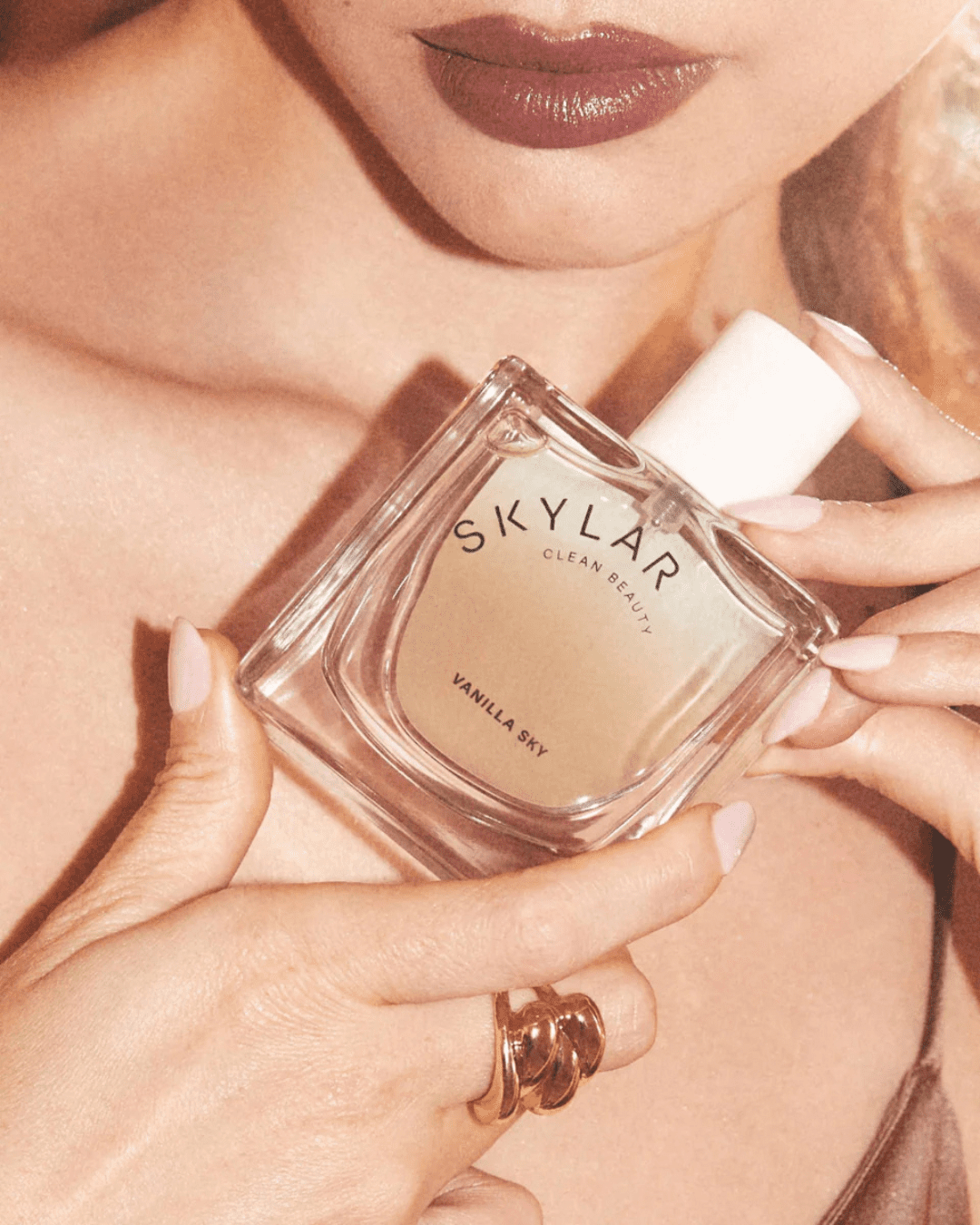 Woman holding Skylar Clean Beauty Vanilla Sky perfume bottle against her chest, wearing a rose gold ring and coffee-colored lipstick, showcasing this cozy ice cream perfume.