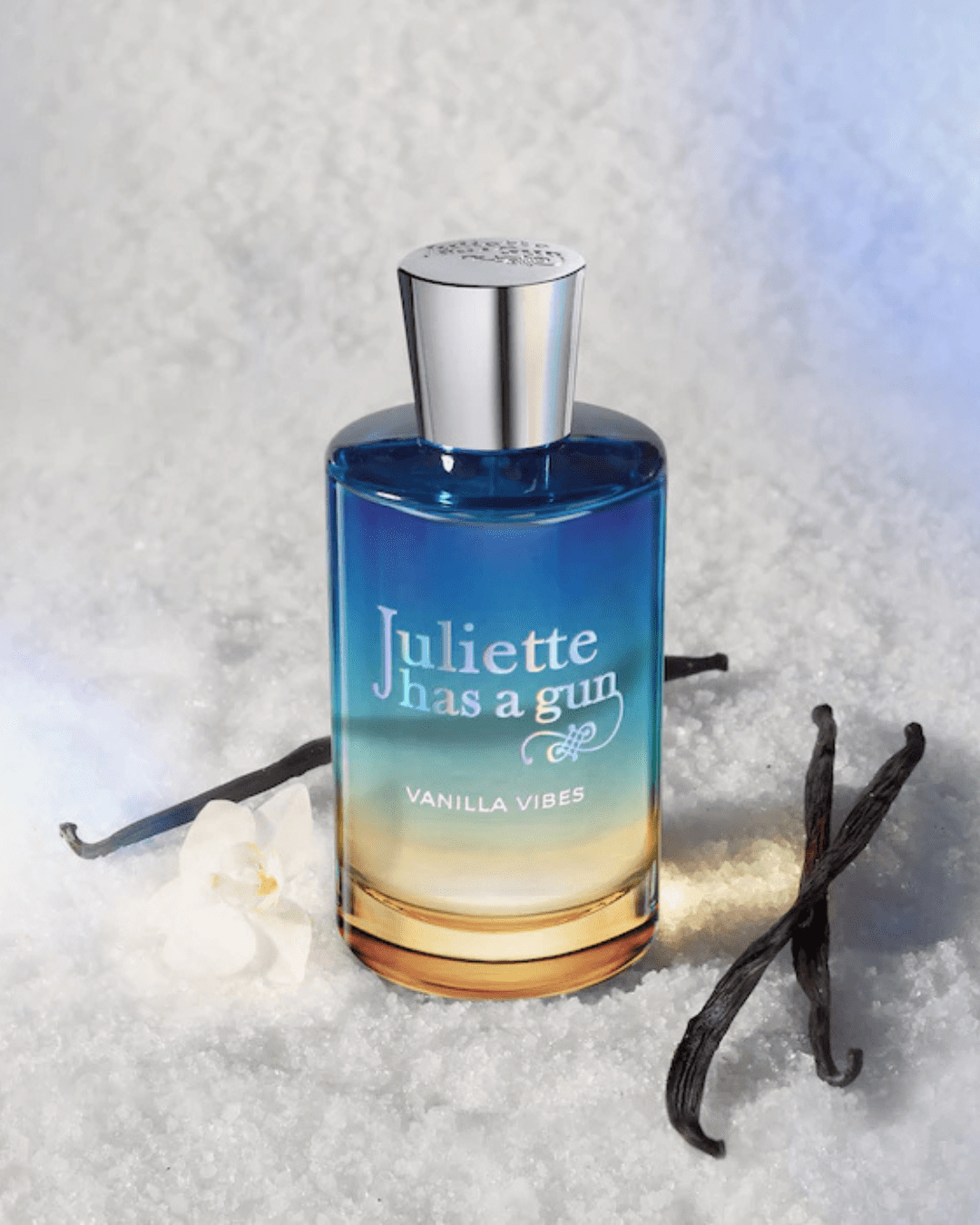 Juliette Has a Gun Vanilla Vibes perfume bottle displayed on a sugary surface with a white flower and scattered vanilla beans, a warm fragrance perfect for layering with ice cream perfumes.