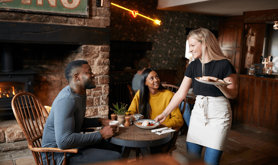 Black man and Indian woman sitting at a restaurant table while a white woman server delivers food to them.