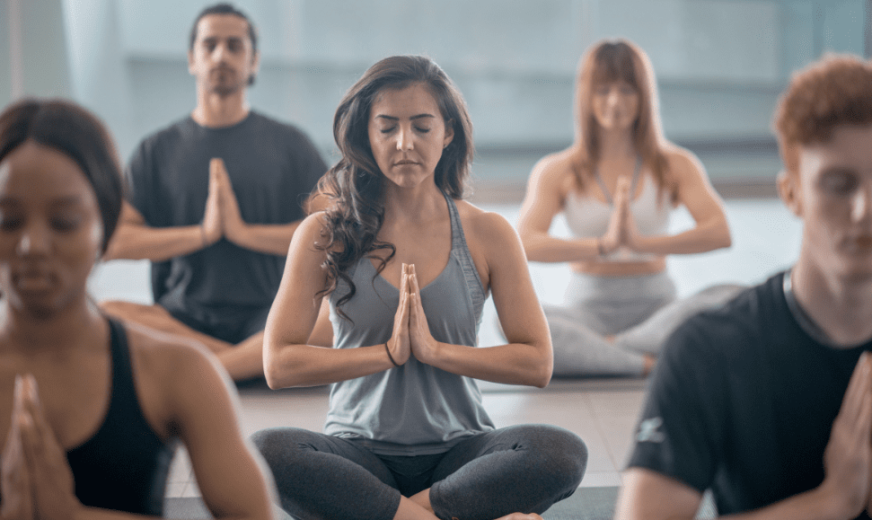 Woman sitting cross-legged in a yoga studio, eyes closed, hands in prayer position, wearing dark gray pants and a light gray spaghetti strap top, hair in curls, surrounded by four other participants slightly out of focus.