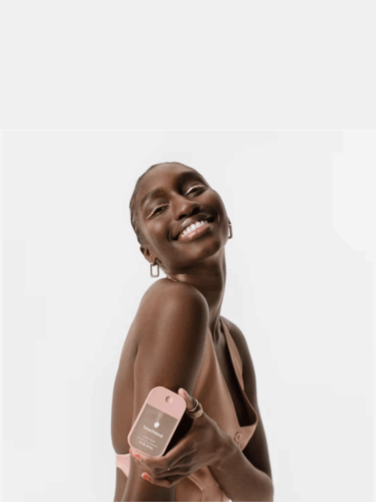 A beautiful black woman poses in a pink vest with no shirt underneath, holding Touchland Glow Miss Rosewater Revitalizing Hand Sanitizer. She crosses one arm over her body, smiling toward the camera with pink eyeliner matching the sanitizer bottle. Her square earrings glint softly against the bright background.