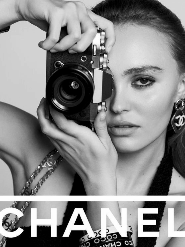 Lily Rose Depp holds a square camera up to her eye at a vertical angle, looking at the camera taking her picture. There is a purse strap made of a Chanel chain with the Chanel logo made into in. She also wears black and white bracelets on her wrist and Chanel earrings with the Chanel logo on them. The black-and-white image features the text "Chanel" across the bottom of the image.