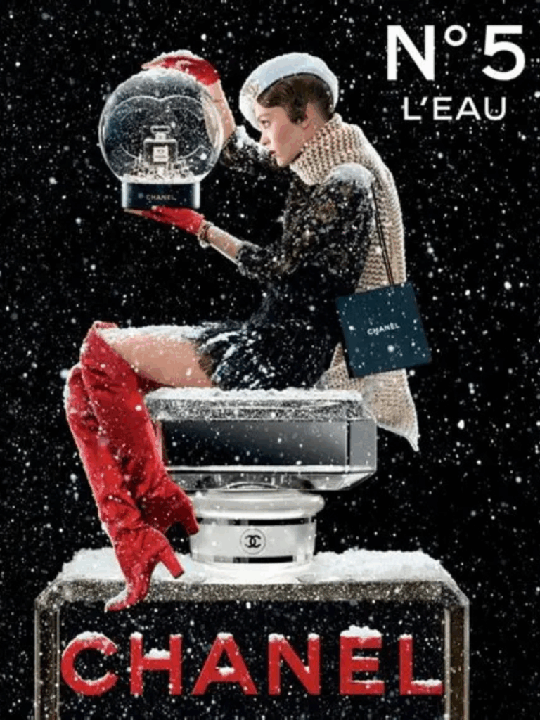 Lily Rose Depp sitting atop a giant Chanel No. 5 L'EAU bottle with "Chanel" in red lettering. She wears red boots that go up to her knees, a black skirt, a black sweatshirt, a thick tan scarf, a dark Chanel bag, and a white Chanel beret with gold Chanel logo. She holds a snow globe containing a Chanel perfume bottle, looking into it, with a black snowy background and snow dusted on the bottle she's sitting on.