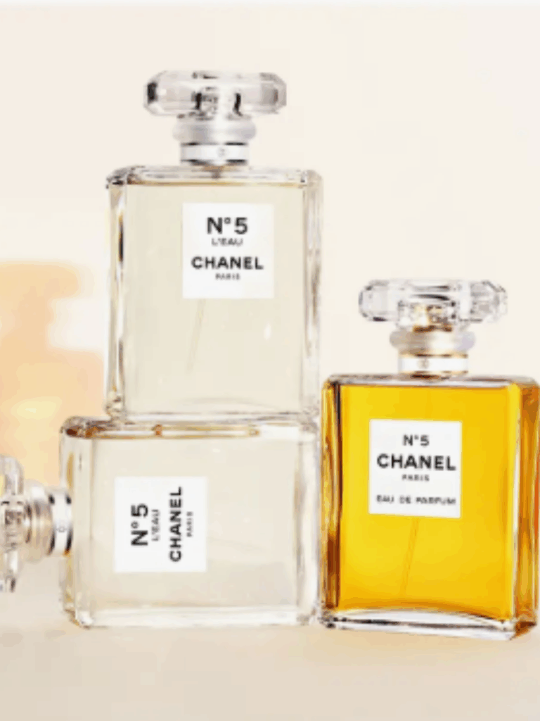 Cream-colored background with two Chanel No. 5 L'EAU bottles, one lying on its side and the other atop it, positioned next to the original Chanel No. 5 bottle, all displayed clearly on the cream surface.