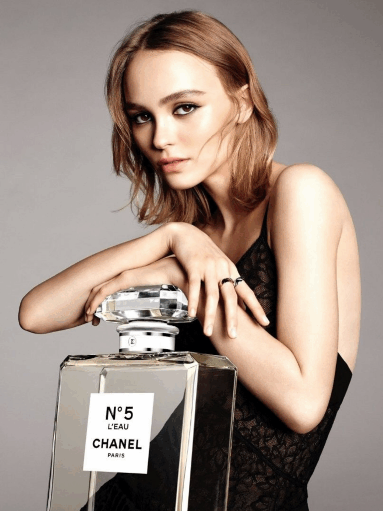 Lily Rose Depp poses with a giant empty Chanel No. 5 perfume bottle, hands crossed on top of the bottle, wearing a black lace see-through dress and looking directly at the camera, creating a striking and elegant fashion image.
